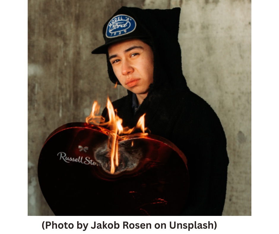 This image shows a young man in a black hoodie and baseball cap holding a heart-shaped box of Valentine's Day candy which is on fire.