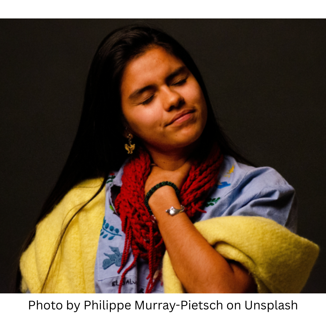 This image represents stress and stored emotions in the body. It shows an indigenous woman with long, dark hair dressed in layers of red, blue and yellow shawls. She has a hand on her neck rubbing it as if it is stiff and uncomfortable.