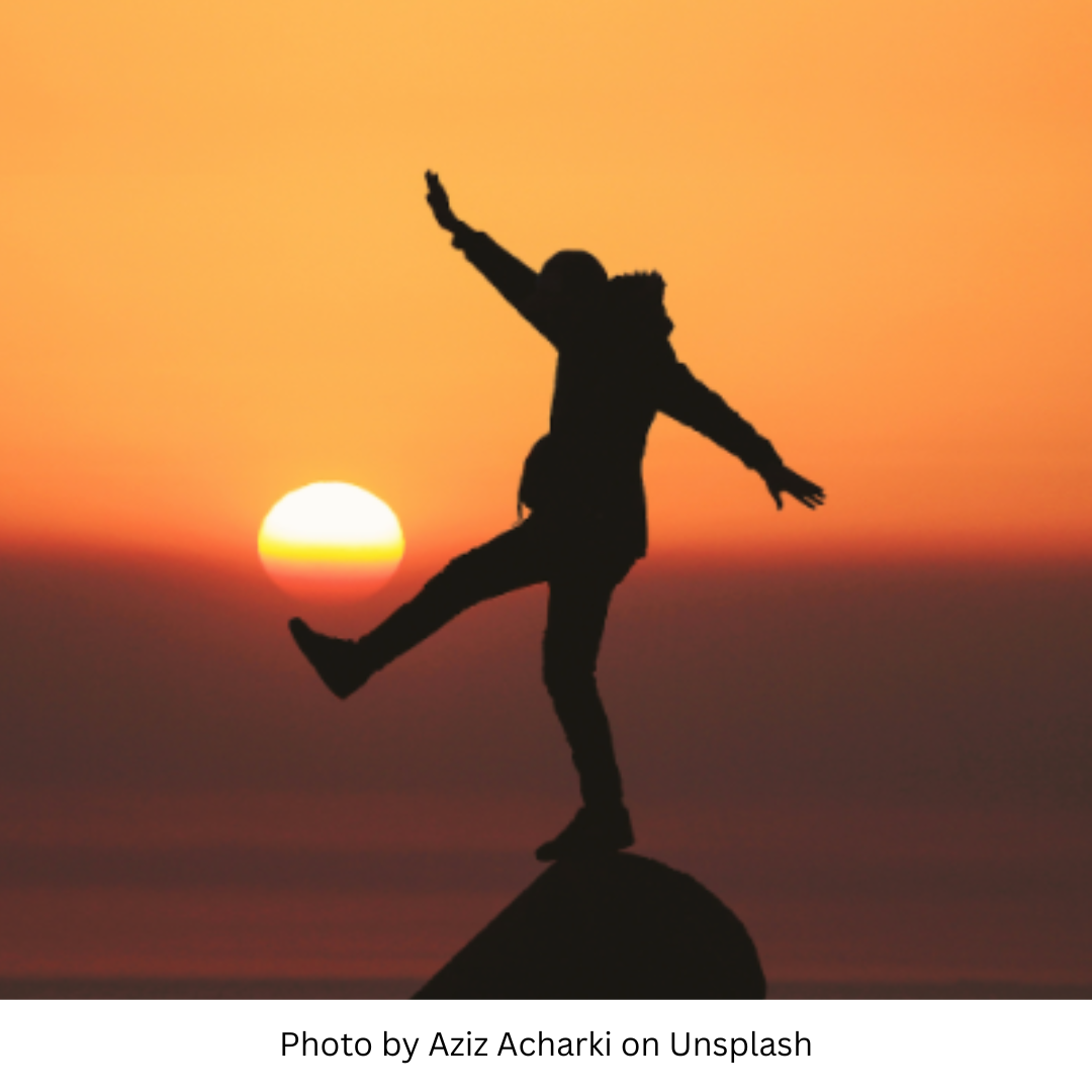 Photo by Aziz Acharki on Upsplash. This image represents the heat and energy of the Spring Equinox by showing a silhouette of a person balancing on one foot on a pointy rock high above the ground. Their arms are out for balance. Behind them is a sunset in shades of red and orange.