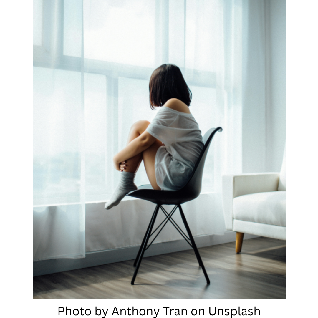 This image shows a petite woman sitting on a tall stool with her knees curled up to her chest. She is looking out the window through a sheer curtain. This represents the discomfort that can be felt when stress causes imbalances in the body's hormones.